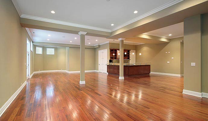 A large, open-concept finished basement featuring polished hardwood floors, recessed lighting, support columns, and a built-in wet bar