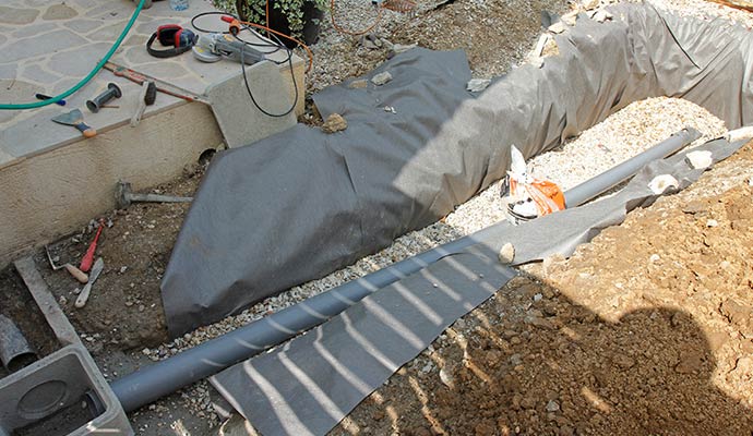 An exterior French drain installation featuring a perforated drainage pipe laid in a gravel trench with landscape fabric and excavated soil.