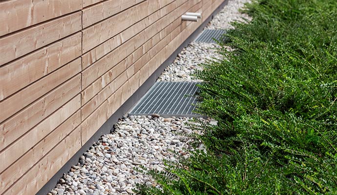 A ground-level view of metal grate basement window well covers installed in a gravel border alongside a modern wood-paneled building exterior.