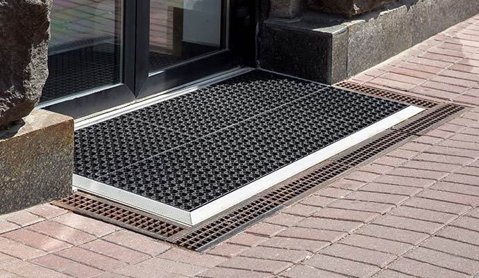 A heavy-duty black rubber scraper mat and metal recessed trench drain system installed at a commercial glass door entrance with brick pavers